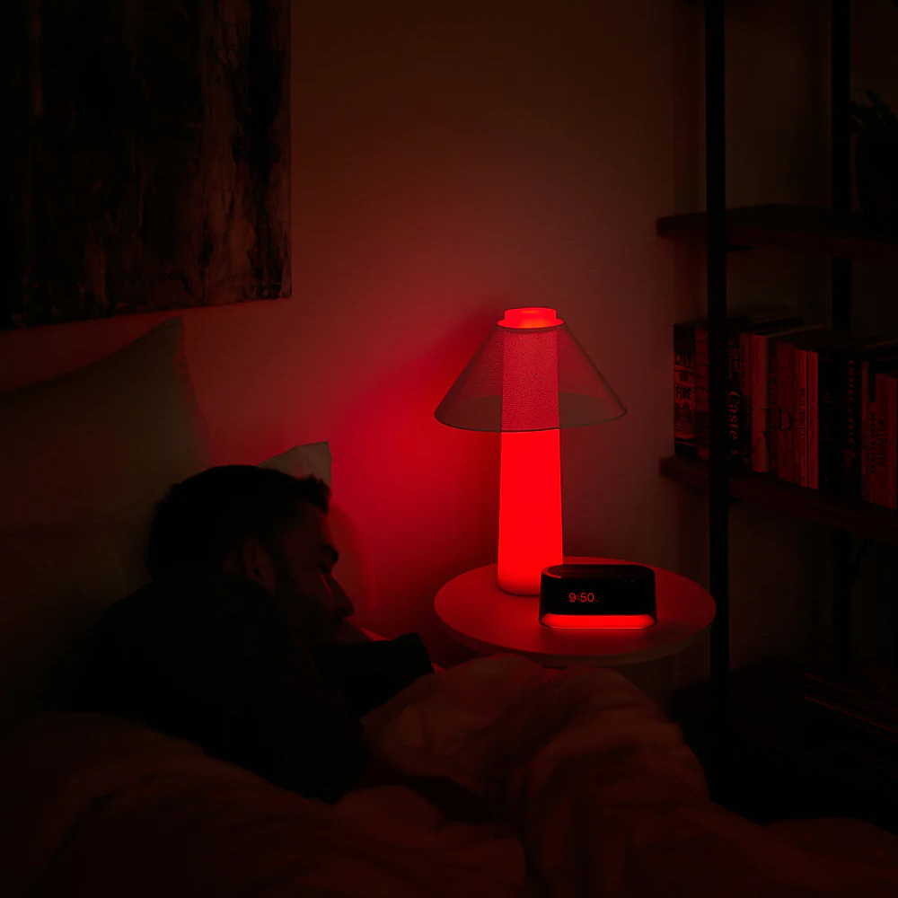 Person sleeping in bed beside a red-lit Loftie lamp and clock reading 9:50, in a dark bedroom.