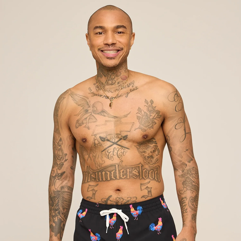 Shirtless man with extensive tattoos smiling, wearing black swim trunks with a colorful rooster print.