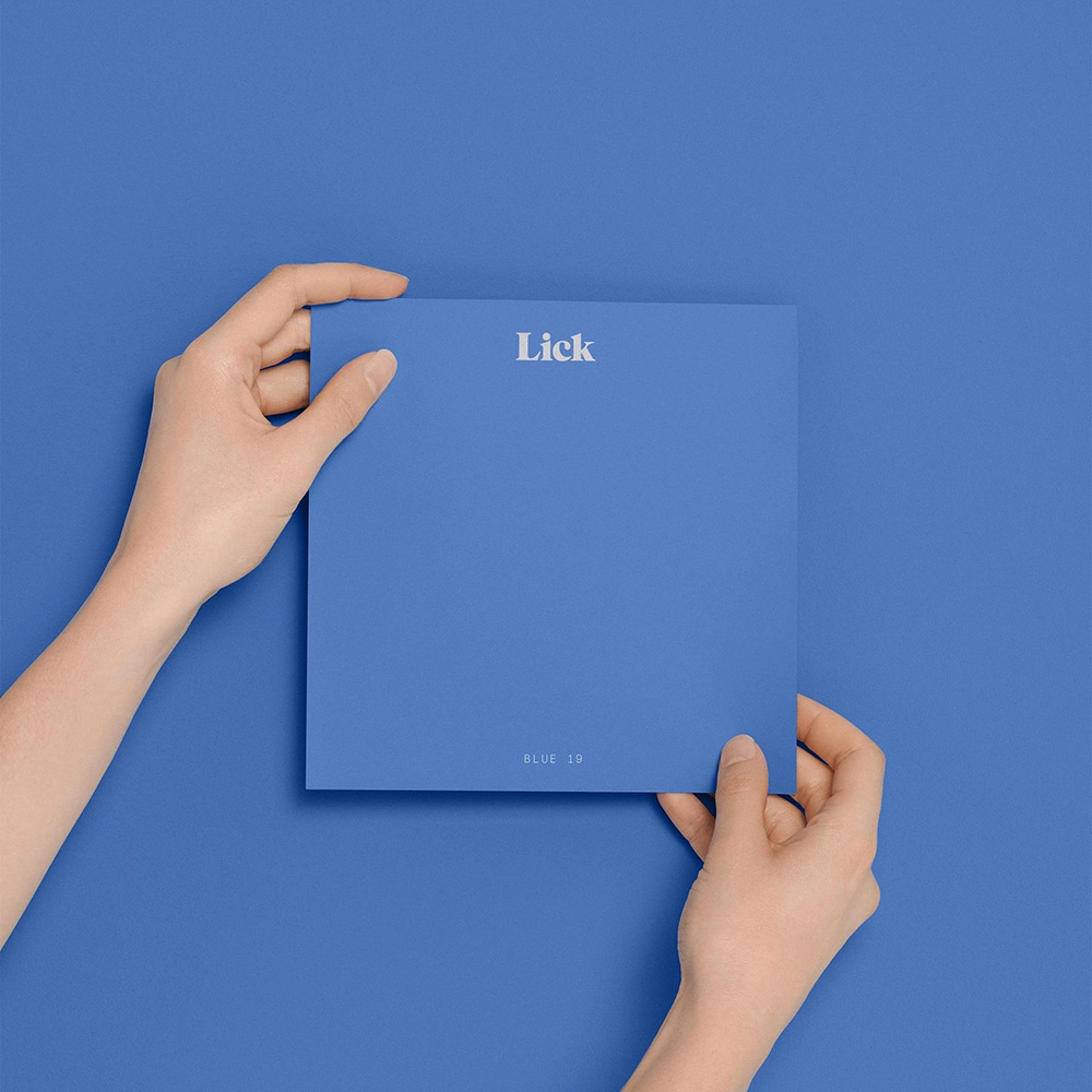 Two hands holding a Lick paint sample card in "Blue 19," matching the blue background.