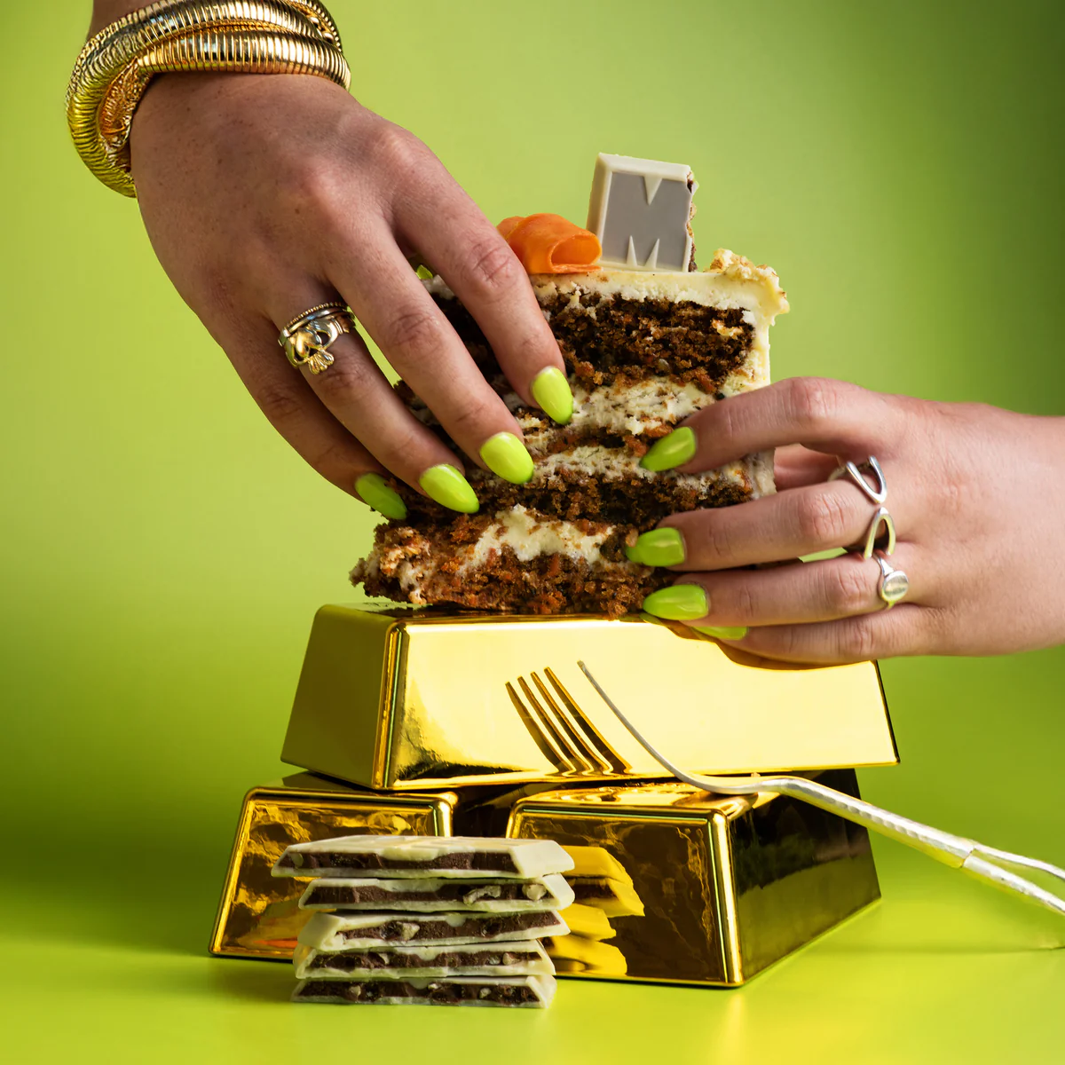 woman's hands with pistacchio green nail polish holding a piece of layered cake on a stack of gold bars