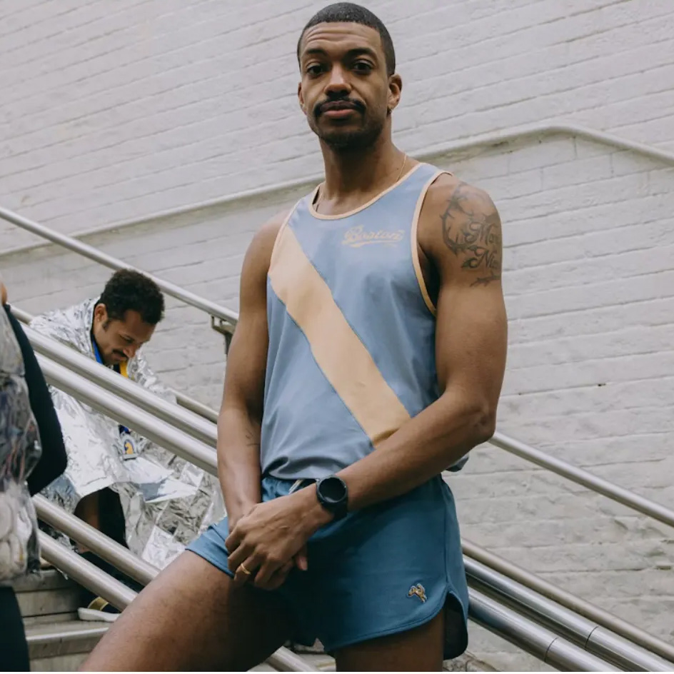 Man in a blue and tan "Boston" tank top and running shorts standing on stairs, with a runner wrapped in a foil blanket in the background.
