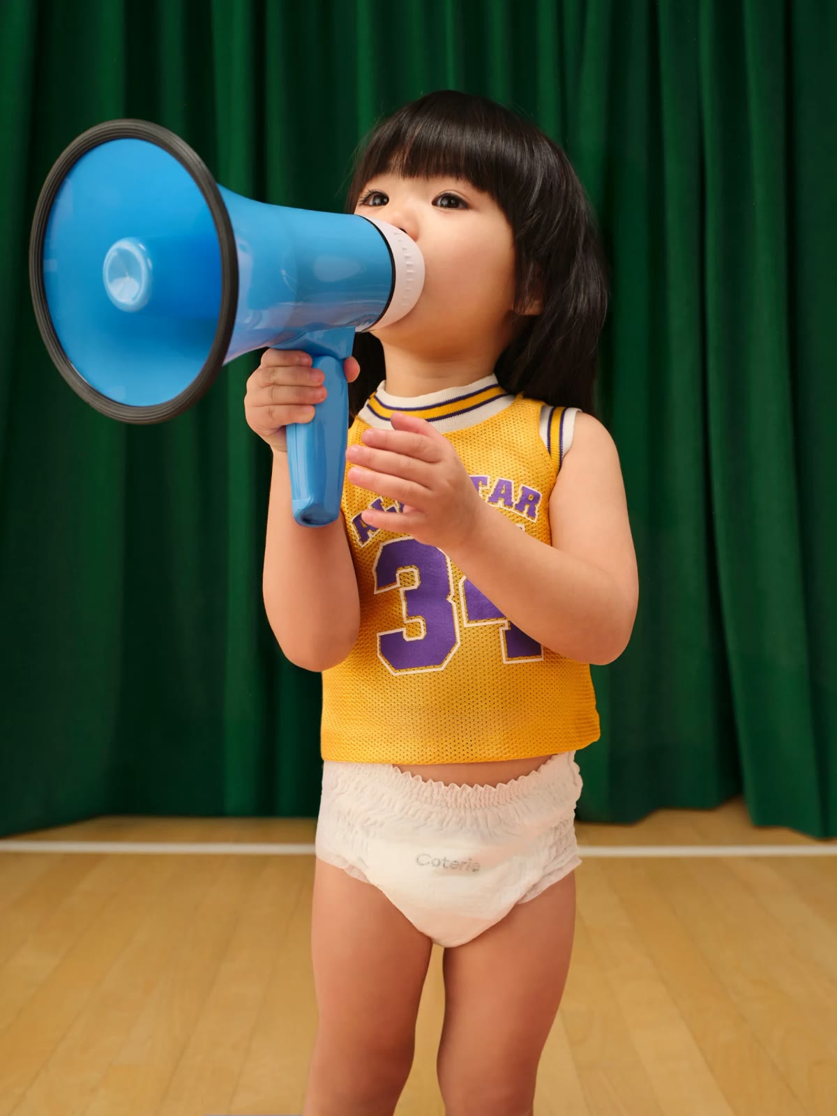 A toddler in a basketball jersey and Coterie diaper speaking into a blue toy megaphone