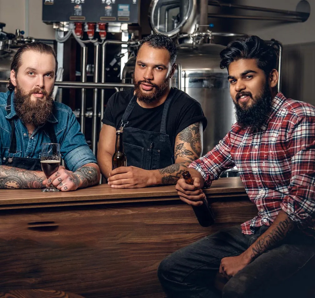 Three bearded brewers with beers and tattoos leaning on a wooden bar in front of brewing equipment.