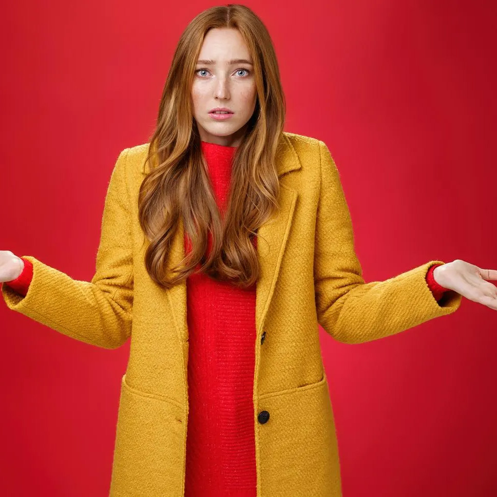 Woman with red hair shrugging with hands raised, wearing a mustard coat over a red sweater against a red background.