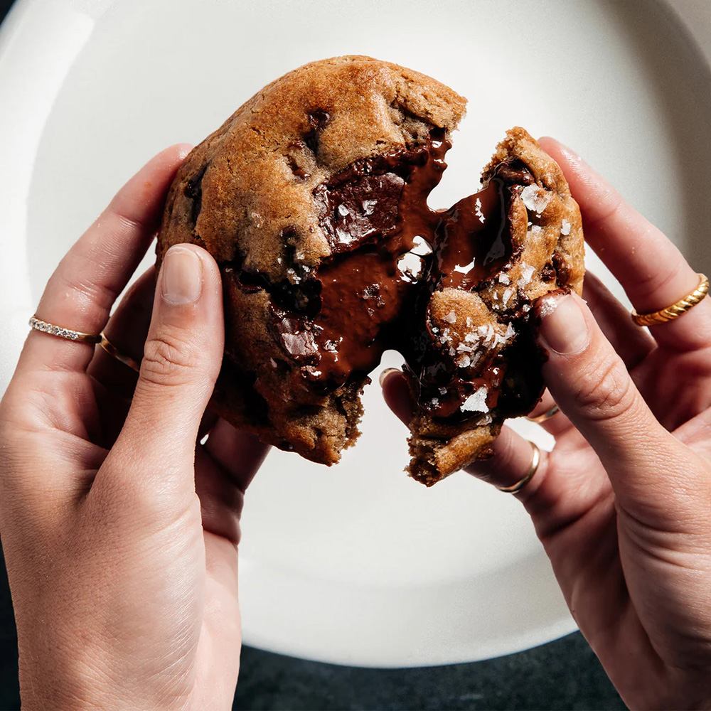 Two hands pulling apart a chocolate chip cookie, revealing melted chocolate and flaky sea salt inside.